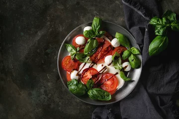 Poster Italian caprese salad with sliced tomatoes, mozzarella cheese, basil, olive oil. Served in vintage metal plate on textile napkin over dark metal background. Top view with space. Rustic style © Natasha Breen