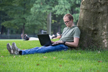 Obraz premium Young man in glasses work on laptop sitting on the grass near the tree in park. Maybe student or hacker businessman