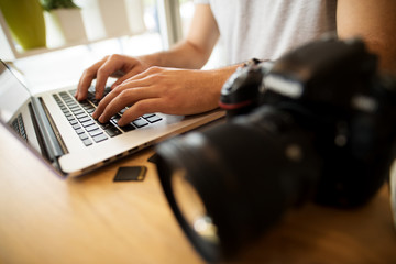 Close up of photographer typing on a laptop with DSLR on the table at home.