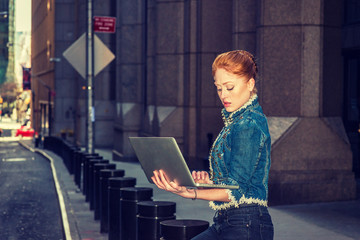 American business woman traveling, working in New York, wearing fashionable jacket, holding laptop computer, sitting on pillar on narrow street, looking down, reading. Instagram filtered effect..
