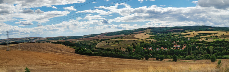 Fototapeta na wymiar A typical natural landscape of Bulgaria: agricultural fields in the hills under a cloudy sky, a village of houses with red roofs. Panoramic view.