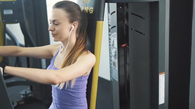 Fit caucasian girl, in purple vest and white headphones, trains arms using chest press machine at the gym
