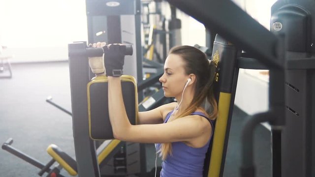 Fit caucasian girl, in purple vest and white headphones, training chest on the pec deck machine at the gym