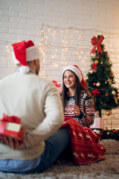 Rear view of young man preparing to give present to his beautiful smiling girlfriend while they sitting on the floor at home for Christmas holidays.