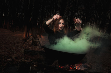 A beautiful girl witch cooking an infernal potion in a cauldron at the stake in a dark forest. The...