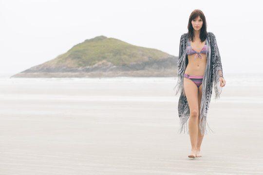 A woman in a pink bikini walking toward the camera on a cloudy beach