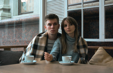 Young nice couple sitting in cafe wrapped in cozy blanket drinking tea. Love story of a girl and a boy. Man and woman embrace one another on a terrace. Sweet couple choosing what to read on a tablet.