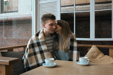 Young nice couple sitting in cafe wrapped in cozy blanket drinking tea. Love story of a girl and a boy. Man and woman embrace one another on a terrace. Sweet couple choosing what to read on a tablet.