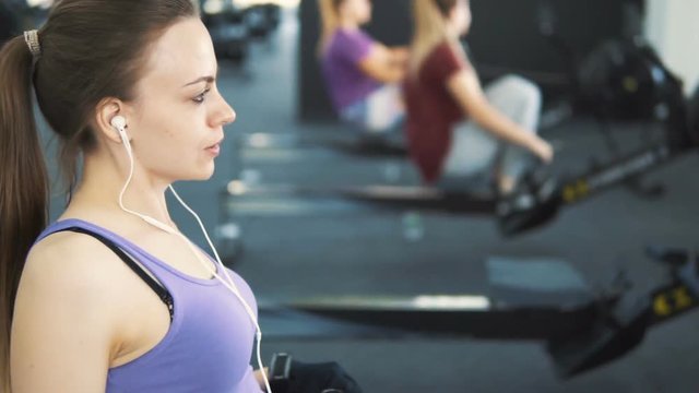 Pretty girl, in purple vest and white headphones, doing rowing workout at the gym