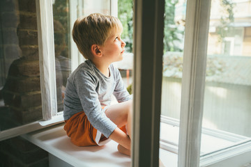Sad preschooler boy sitting on windowshill. A child looks out the window. Vintage color. Childhood,...