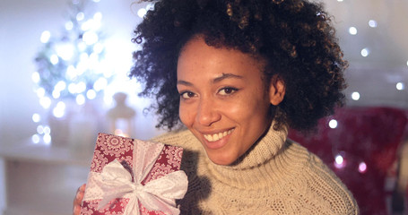 Single beautiful African woman with smile holding Christmas gift wraped with white bow. Includes Christmas tree in background.