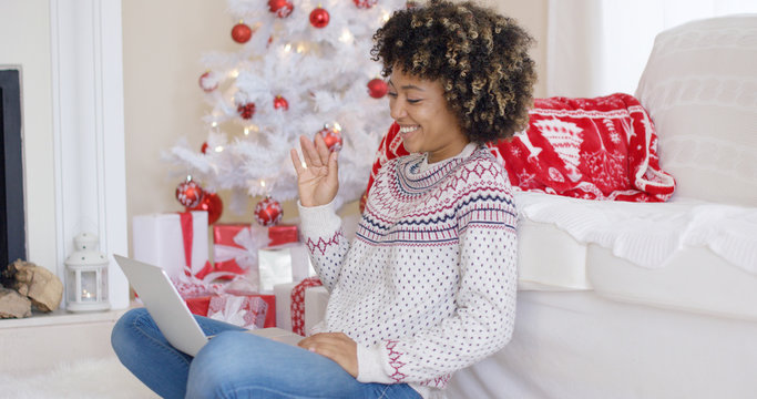 Young woman having a video chat on her laptop waving at the computer as she sits in a red and white themed Christmas living room