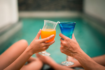 Hands holding juice glasses by the pool summer time