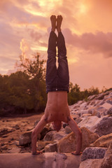Young Caucasian man is hand balancing on the rock at colorful sunset on background