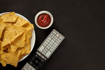 Nachos chips with sauce and TV remote on a black table, top view