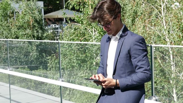 Handsome eleganty young business man wearing jacket standing, taking photo with tablet computer and then sending it to someone, outdoor