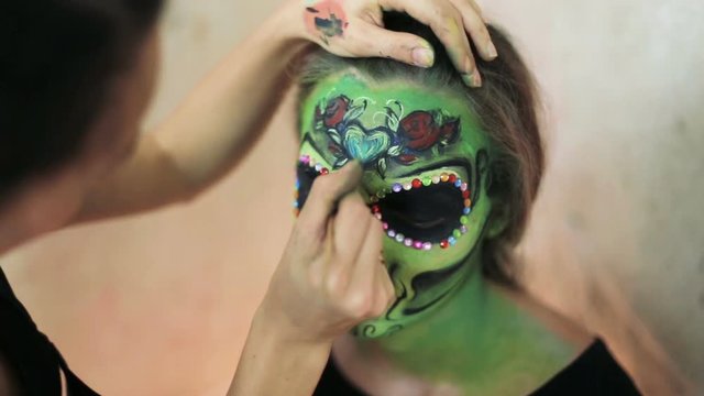 woman doing aquagrim face art on halloween make-up with her hands tassels green scary glamorous skeleton. Mexican Princess Sugar Skull.