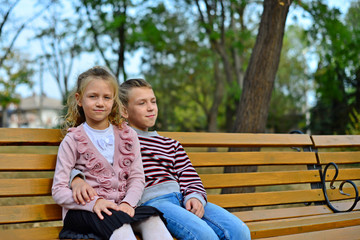 Brother and sister cuddling and sitting on a bench in a park on autumn day. Little girl and boy hugging