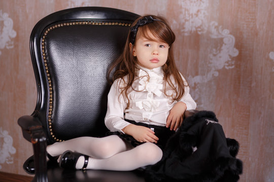 Young brunette dolly lady girl stylish dressed like a boss in white official shirt black skirt posing sitting in studio on black leather chair with pout lips and pink cheeks.