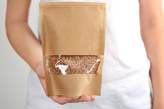 Woman holding paper package with raw buckwheat on light background