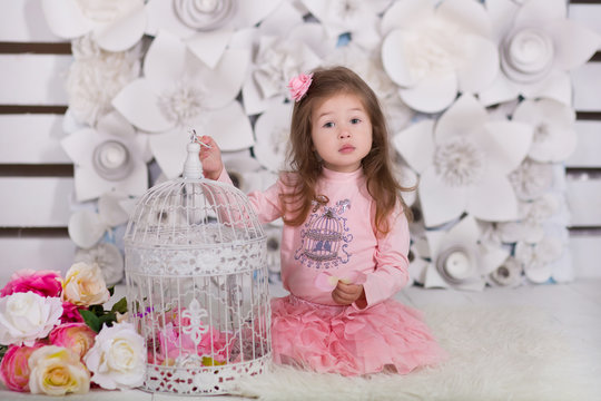 doll girl with brunnete hair stylish dressed in pink dress pinky skirt shirt happyly smiling posing to camera with white parrot cage in modern showroom studio on fur floor with paper flowers on wall.