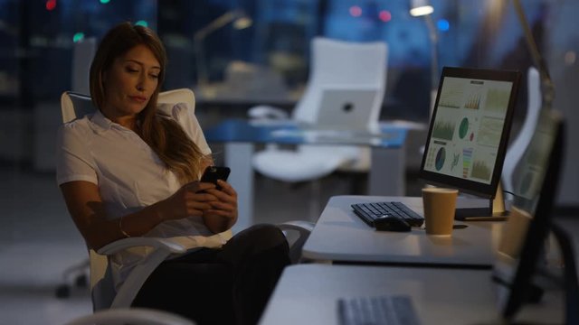  Businesswoman working late in the office, looking at smartphone at her desk