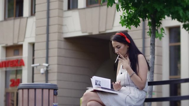 A girl reads a book in the park while smoking and starts yawning.