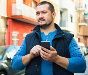 Man in blue stands on street and looks at his mobile phone