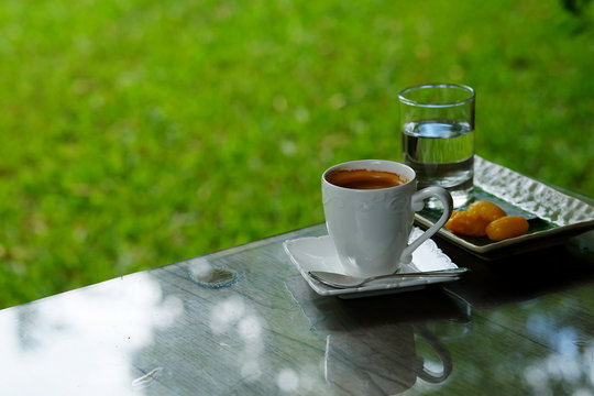 hot black coffee or hot American coffee in white cup Served with Met Khanoon (Golden Jackfruit Seeds Thai Mung Bean Marzipan and Egg Yolk Dessert) green lawn garden.have some space for write wording