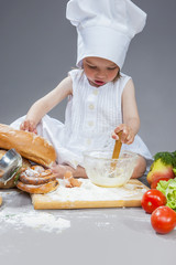 Cooking Ideas. Caucasian Little Girl In Cook Uniform Working With Whisk and Kitchen Glassware In Studio Environment. With Vegetables and Fruits on Background.