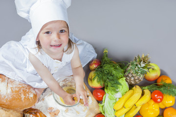 Cooking Ideas. Caucasian Little Girl In Cook Uniform Working With Whisk and Kitchen Glassware In Studio Environment. With Vegetables and Fruits on Background.High Angle View.