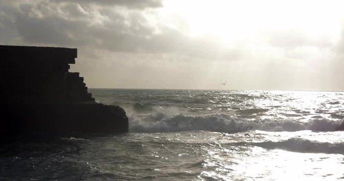 Sea sparkling in the sun rays striking through the clouds. Waves crushing the ancient walls of Acre city, Israel. Sea-gulls flying over the water