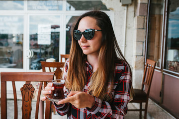 Beautiful young girl with sunglasses on head smiling sitting at table in an outdoor cafe and drinking tea from glass beaker