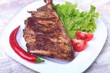 Pork ribs on grill and chili pepper with tomato, lettuce leaves on white plate