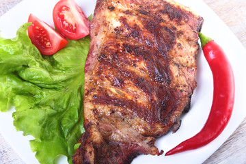 Pork ribs on grill and chili pepper with tomato, lettuce leaves on white plate