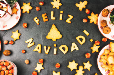 FELIZ NAVIDAD COOKIES. Words Merry Christmas en Spanish with baked cookies, Christmas decoration and nuts on black slate background. Christmas card for hispanic countries top view
