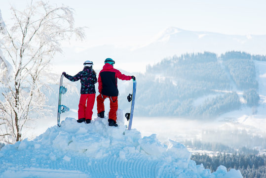 Couple snowboarders - man and woman enjoy the snow-white scenery of mountains and forests of the Carpathians from the height of the mountain top at winter ski resort. Back view