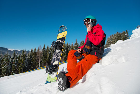 Full length shot of a snowboarder resting sitting on the slope, talking on the phone at ski resort on a beautiful sunny winter day in the mountains