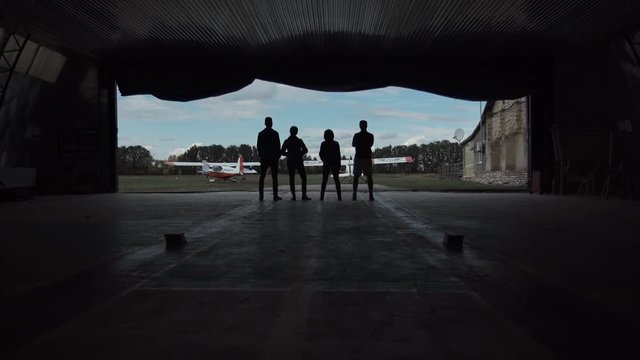 Four people standing in an aircraft hangar silhouetted against the sky watching two small aircraft outside in a low angle view