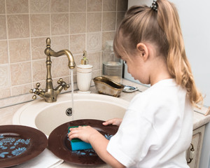 A little cute little girl, four years old, washes the dishes in the kitchen. Water runs in the sink. The child plays and helps mom. 