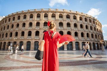 Woman in red dress with spanish hand fan stnading back in front of the bullring amphitheatre in...