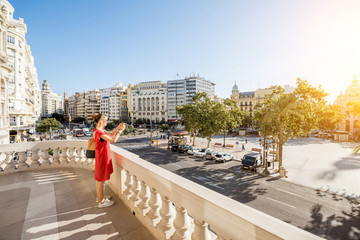 Young woman tourist in red dress photographing Ayuntamiento square from the terrace of city hall building in Valencia during the morning light in Spain