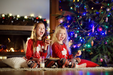 Happy little sisters having milk and cookies by a fireplace in a cozy dark living room on Christmas eve