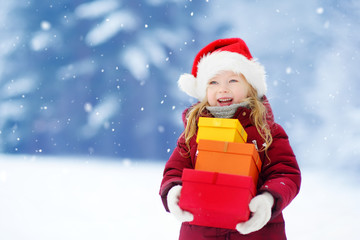 Adorable little girl wearing Santa hat holding a pile of Christmas gifts on beautiful winter day