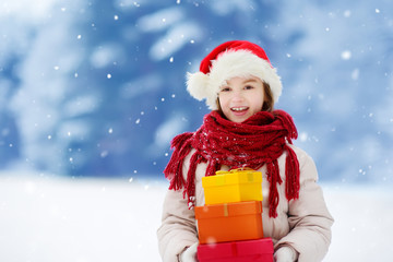 Adorable little girl wearing Santa hat holding a pile of Christmas gifts on beautiful winter day