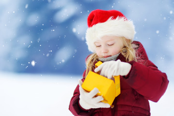 Adorable little girl wearing Santa hat holding Christmas gift on beautiful winter day