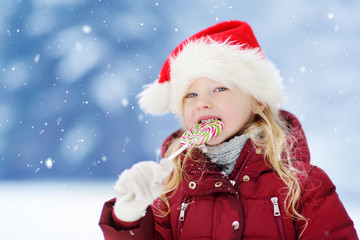 Adorable little girl wearing Santa hat having huge striped Christmas lollipop on beautiful winter day