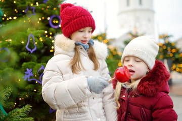 Two adorable little sisters eating red apples covered with sugar icing on traditional Christmas market