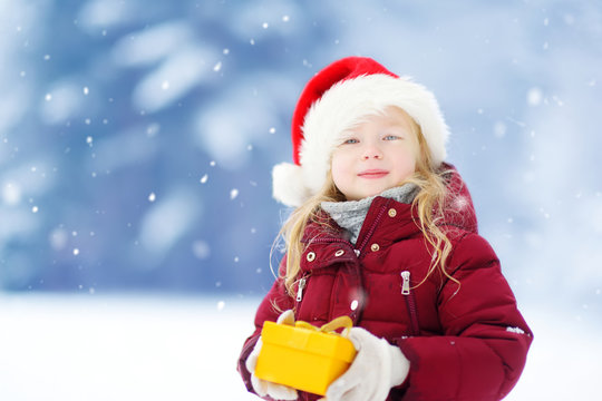 Adorable little girl wearing Santa hat holding Christmas gift on beautiful winter day