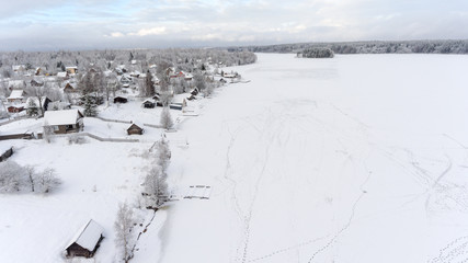 Fototapeta na wymiar Footprints are on ice of frozen lake near winter shore. Russian remote village is in evergreen forests. Aerial view. Russia